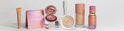 Auf einer weißen Oberfläche werden verschiedene Make-up-Produkte von LH cosmetics präsentiert, darunter Rouge, Highlighter, Foundation und Lipgloss.