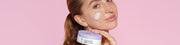 Frau mit frischer Haut trägt Creme auf ihre Wange auf und hält eine Feuchtigkeitscreme von Geek&Gorgeous in der Hand vor einem rosa Hintergrund.