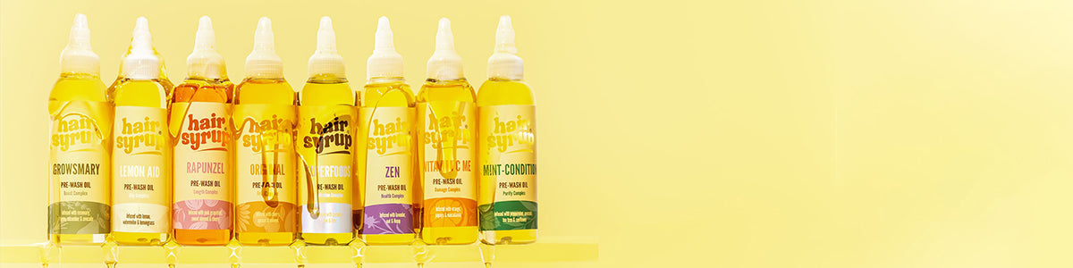 Mehrere bunte Hair-Syrup-Pre-Wash-Öle stehen nebeneinander vor gelbem Hintergrund in einer Reihe.