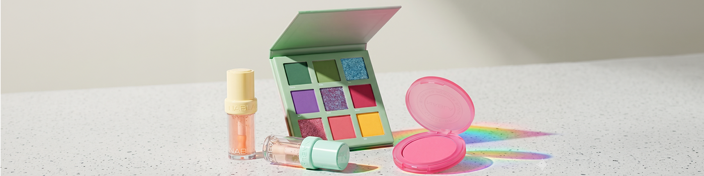 Pastellfarbene Make-up-Artikel, darunter eine Lidschattenpalette, Rouge, Gloss und Highlighter, auf einer gesprenkelten Oberfläche.
