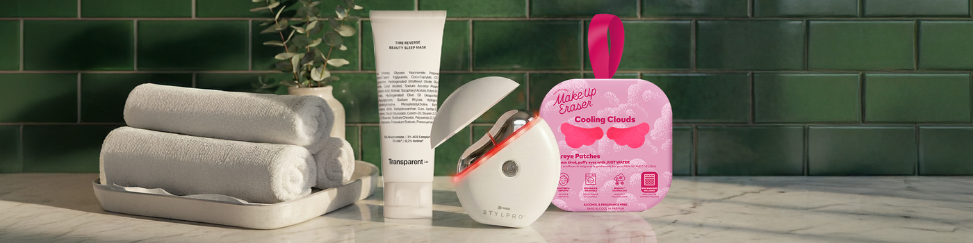 Drei gerollte Handtücher, Hautpflegeprodukte und eine rosafarbene Schönheitsmaske auf einem Badezimmertisch mit grüner Kachelwand.