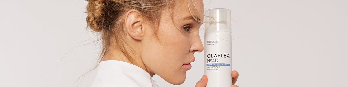 Frau mit blondem Haar im Dutt hält eine Flasche Haarpflegeprodukt Olaplex Nr. 0 nah an ihr Gesicht.