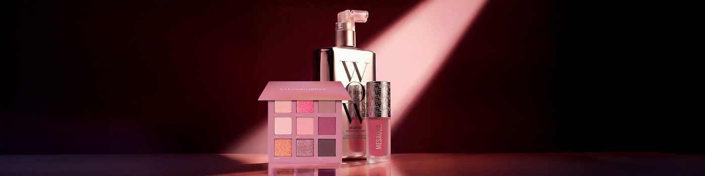 Rosa Lidschattenpalette, Lipgloss, Wimperntusche und Parfümflasche auf einer glänzenden Oberfläche unter einem Scheinwerferlicht.