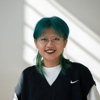 Porträt von Kaiyue mit blauen Haaren, braunen Augen, silberner Brille und schwarz-weißem Oberteil vor hellem Hintergrund.