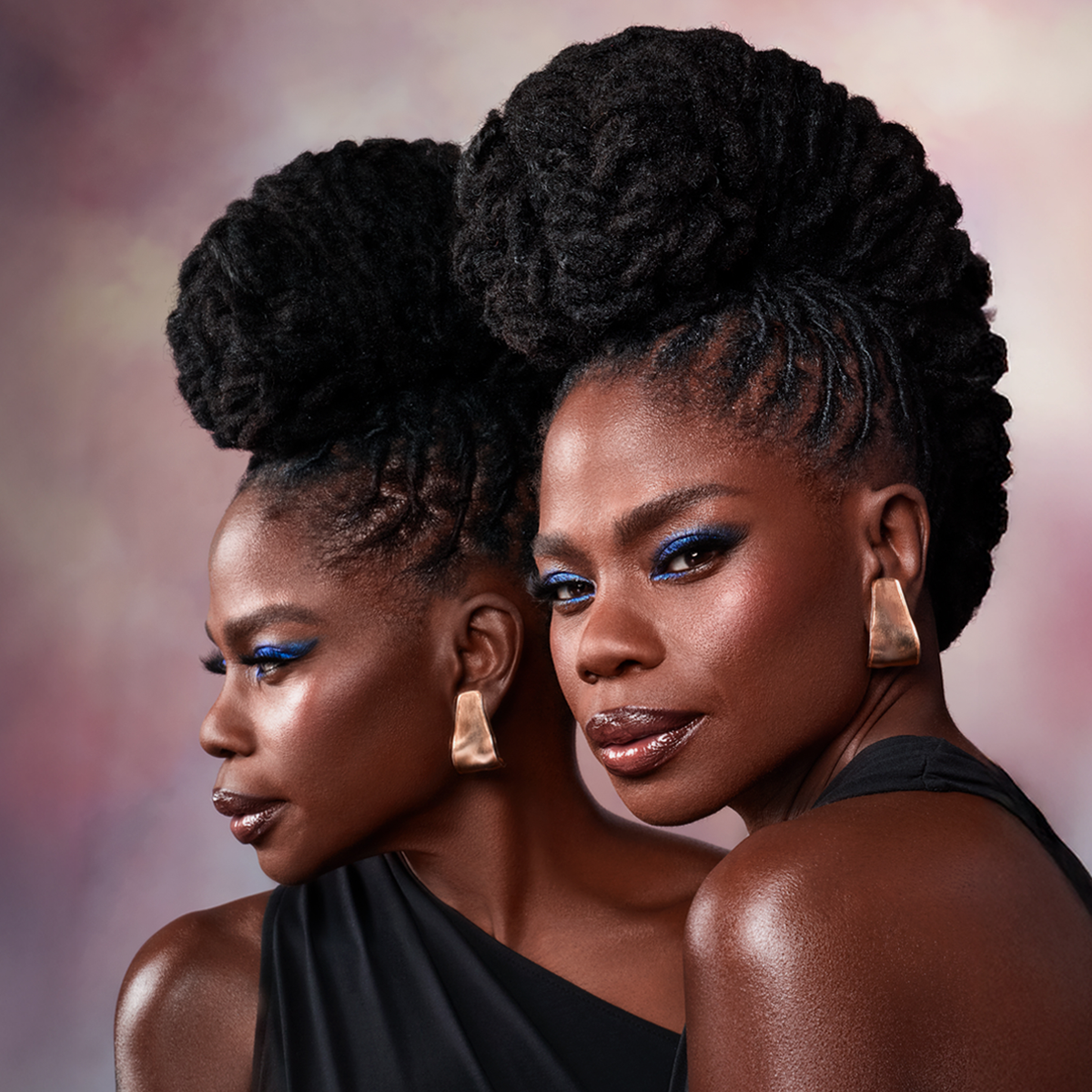 Make-up Stylistin Danessa Myricks mit kräftigem blauen Augen-Make-up, goldenen Ohrringen und aufwendiger Flachtfrisur posierend vor einem Spiegel und zartrosa Hintergrund.