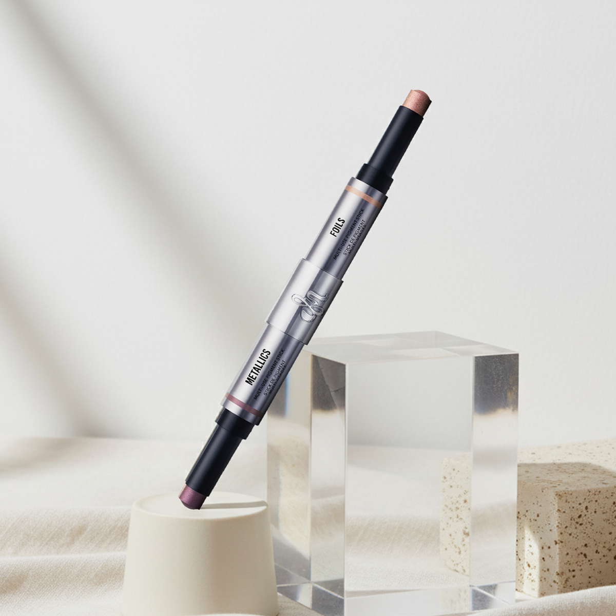 Der Danessa Myricks Beauty Duo Multi-Use Pigment Stick aus dem Signature by Danessa Bundle mit Rosa- und Lilatönen auf Stein- und Glaspodesten vor weißem Hintergrund weißen Fläche. 