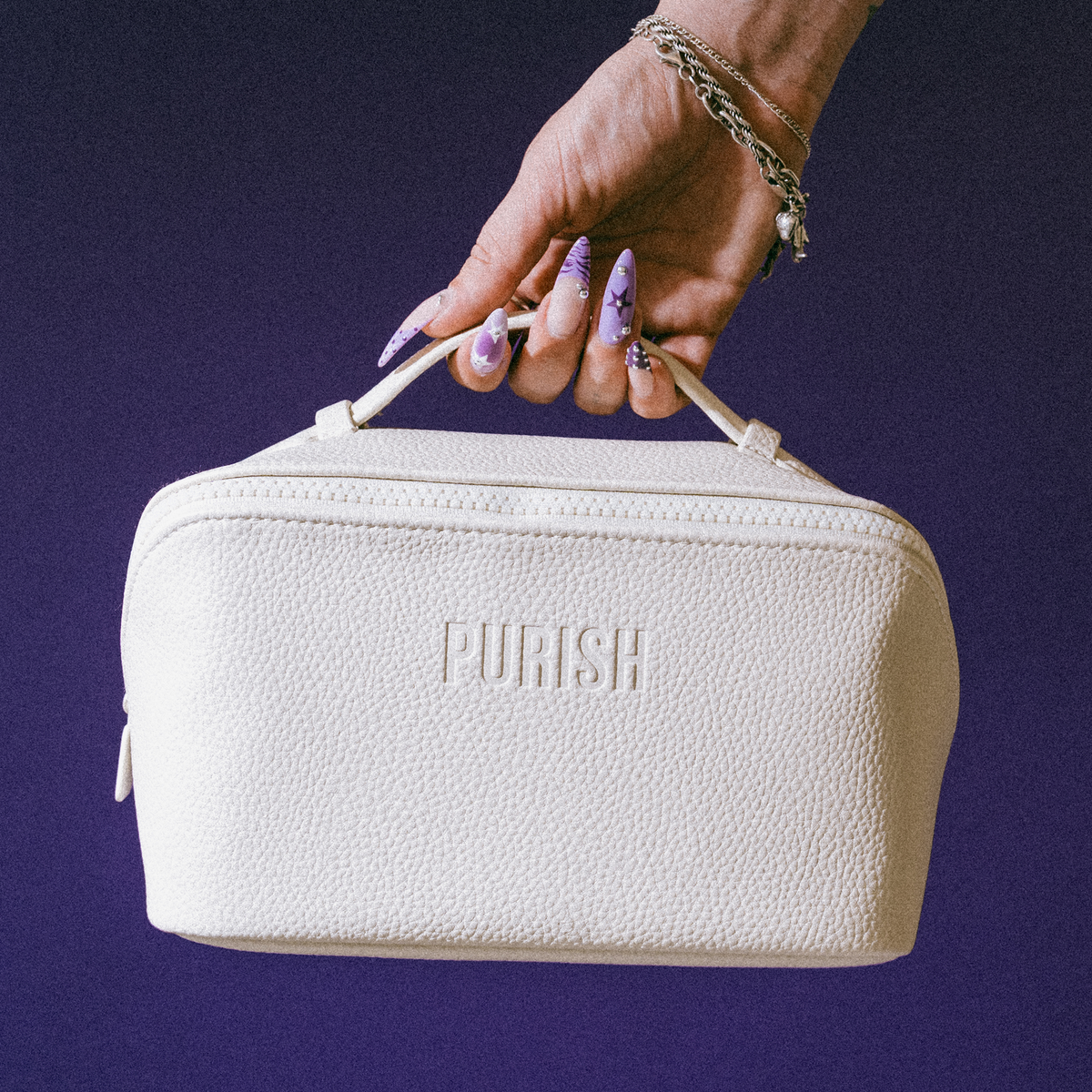 Eine Hand mit langen, verzierten Nägeln hält die PURISH Reisetasche – Creme vor einem violetten Hintergrund.