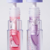 Nahaufnahme von zwei Flaschen „BLONDESISTER 3 IN 1 CUTE LIPPY LIP OIL“ mit roter und violetter Flüssigkeit vor einem hellen Hintergrund.
