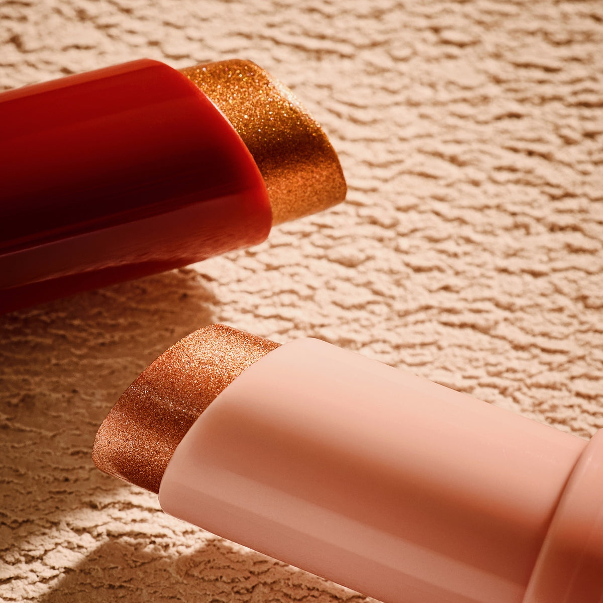 Zwei MIDNIGHT SUN Jelly Glow Sticks von Catrice Cosmetics in roter und rosafarbener Hülle liegen auf einer strukturierten beigen Oberfläche.