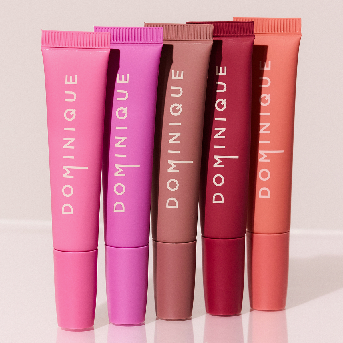 Fünf Tuben Dominique Cosmetics Juicy Lip Balm in verschiedenen Rosa-, Mauve- und Korallentönen, aufrecht in einer Reihe stehend.