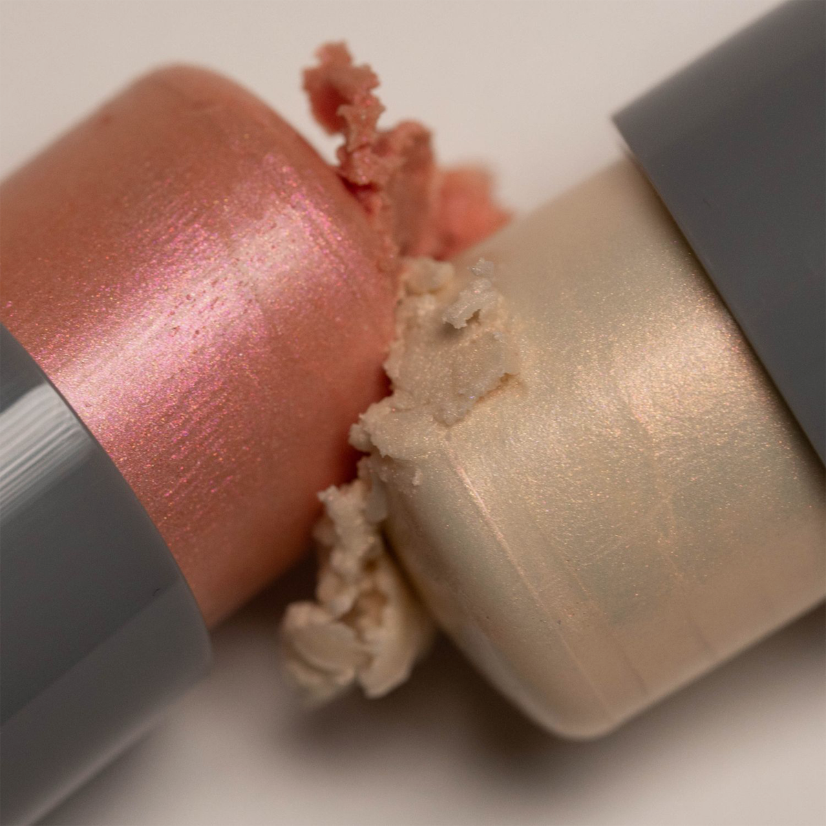 Zwei Artistick Highlighter von LH Cosmetics, einer rosa und einer beige, mit schimmernden, strukturierten Enden, die zusammengedrückt werden.