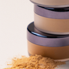 Drei gestapelte LH Cosmetics Infinity Filter Puder mit losem Puder davor auf einer weißen Fläche.