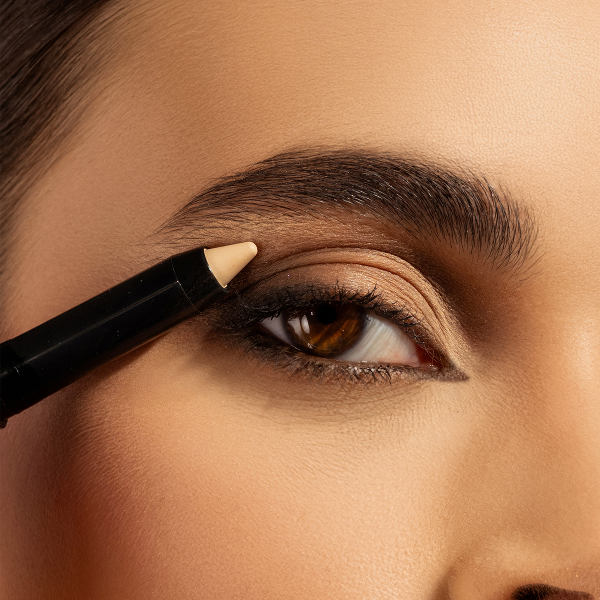 Nahaufnahme eines braunen Auges mit schwarzem Eyeliner-Look, betont durch einen beigefarbenen Lidschattenstift, der am Oberlid aufgetragen wird.