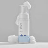 Runde weiße Flasche des Round Lab 1025 Dokdo Bubble Foam mit Pumpspender, umgeben von weichem, weißem Schaum. Schlichtes Design mit blauer Wasser-Illustration.