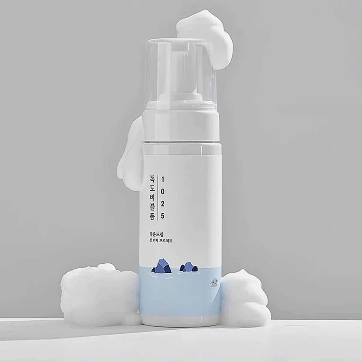 Runde weiße Flasche des Round Lab 1025 Dokdo Bubble Foam mit Pumpspender, umgeben von weichem, weißem Schaum. Schlichtes Design mit blauer Wasser-Illustration.