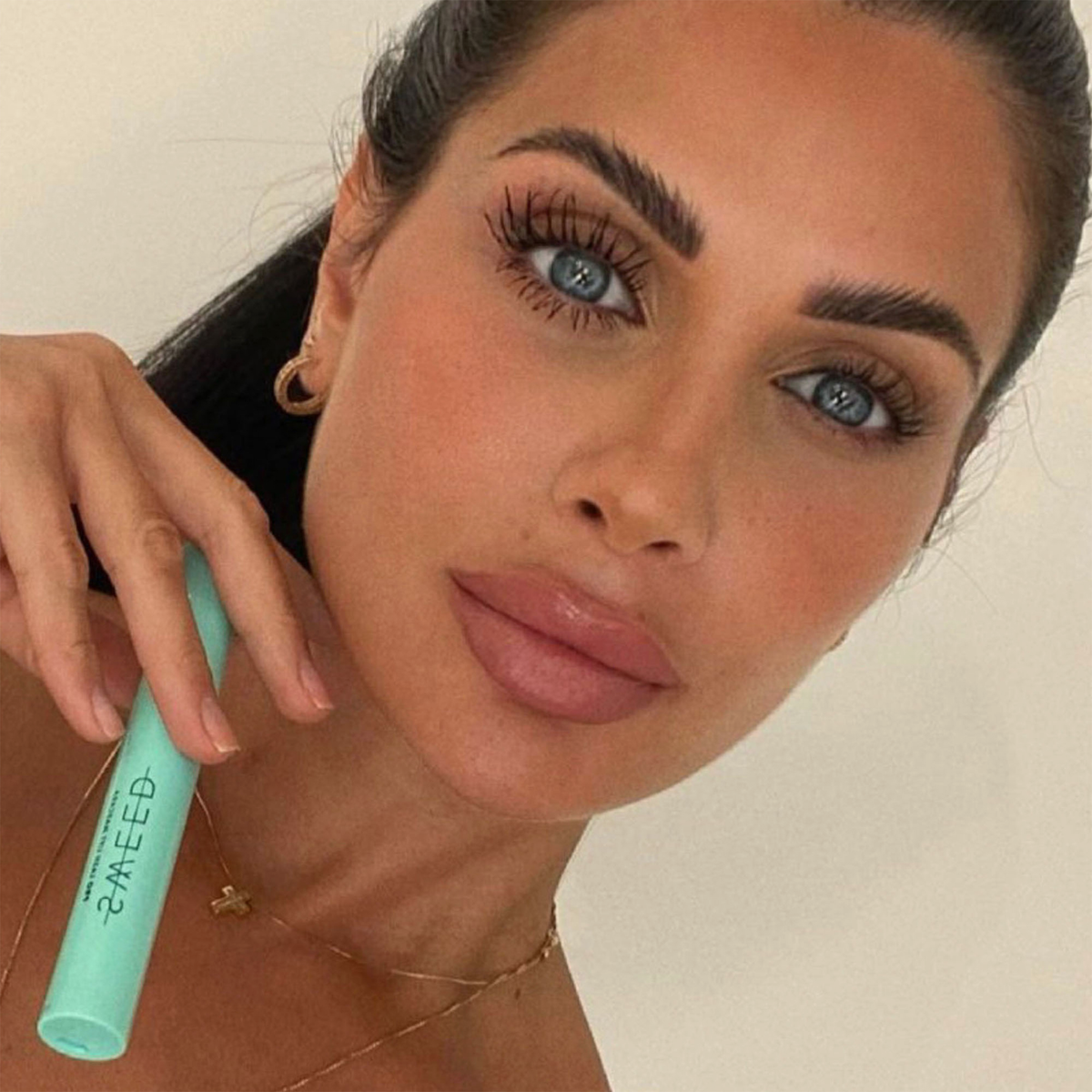 Eine Frau mit langen Wimpern hält die mintgrüne SWEED Lash Lift Mascara in der Hand, trägt Goldschmuck und ist natürlich geschminkt, vor einem beigen Hintergrund.