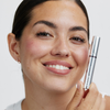 Eine Frau lächelt, während sie eine silberne Tube XLASH Brow Booster in der Hand hält.