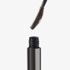 Eine Nahaufnahme der braunen Mascara-Bürste des „XLASH Drama Tinted Primer“ über der geöffneten Metalltube.