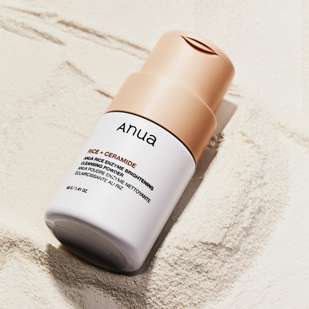 Eine Flasche Rice Enzyme Brightening Cleansing Powder steht auf einer strukturierten, sandigen Oberfläche.