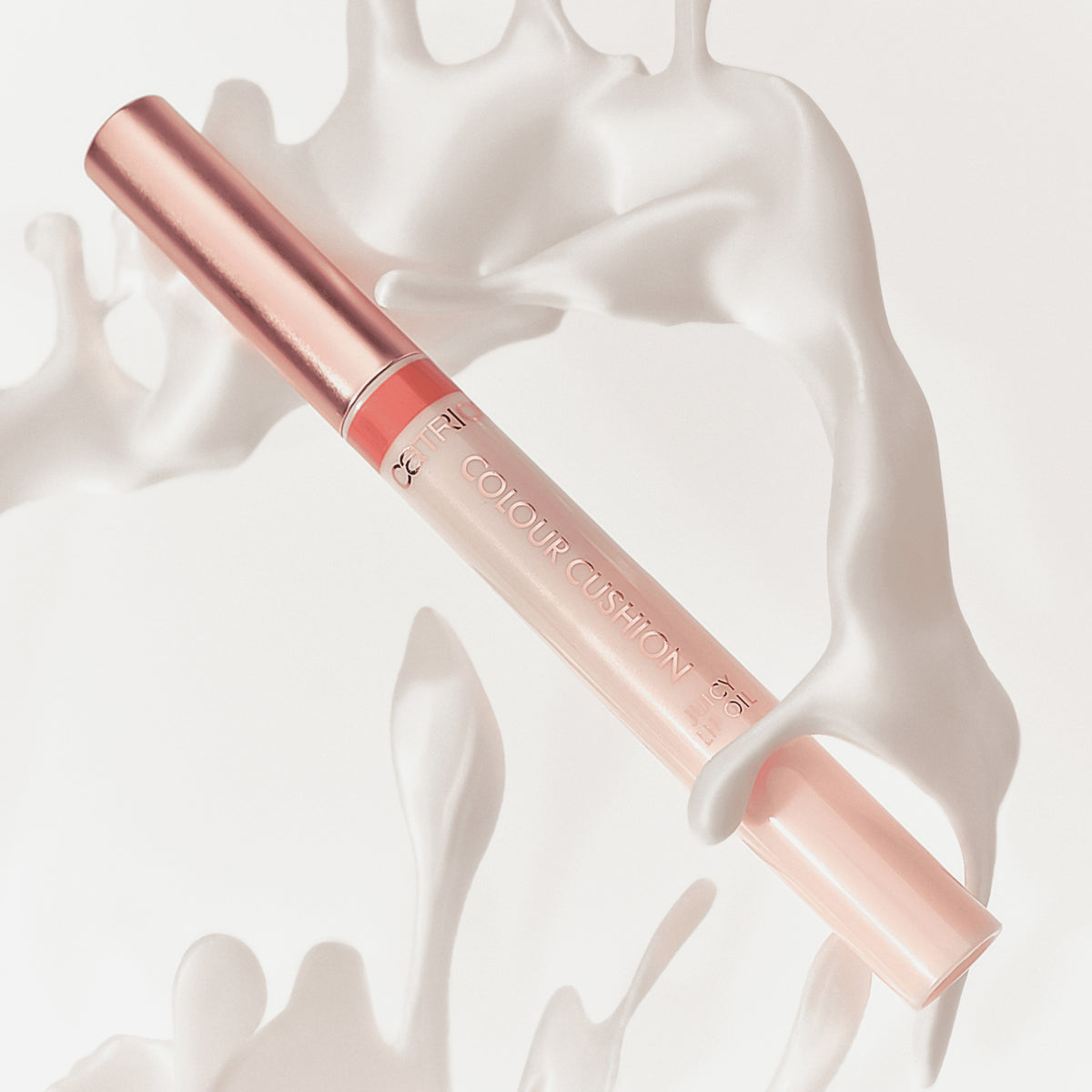 Das Colour Cushion Juicy Lip Oil von Catrice Cosmetics in einer rosafarbenen und roségoldenen Tube schwimmt inmitten von cremeweißen Flüssigkeitsspritzern.