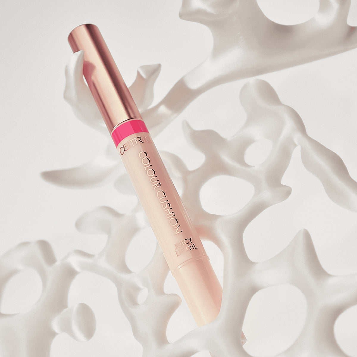Ein Catrice Colour Cushion Juicy Lip Oil in Rosé und Pink liegt dekorativ in weißem, organisch geformtem Dekor.