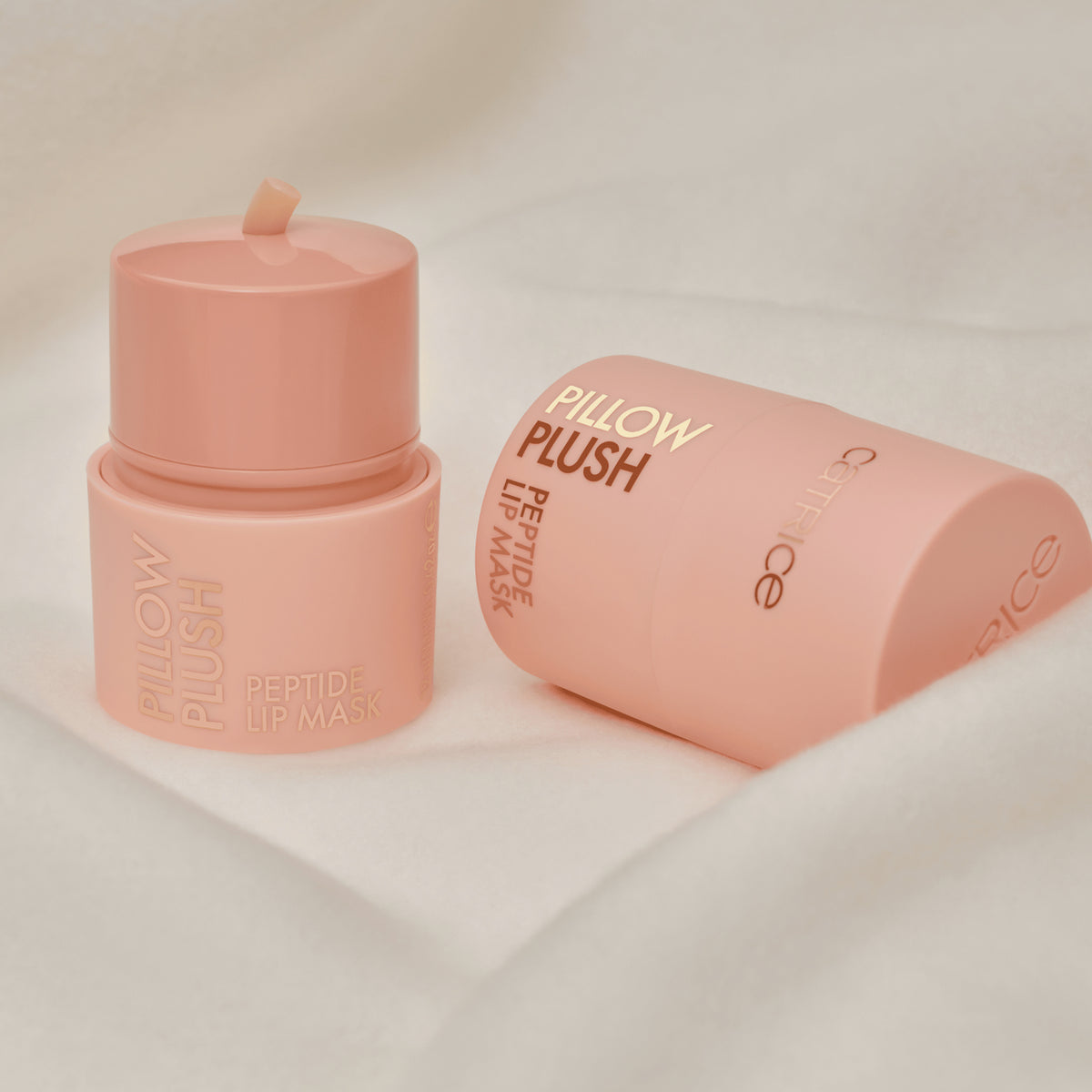 Zwei rosafarbene Behälter von Catrice Cosmetics Pillow Plush Peptide Lip Mask 010 sind auf einem weichen, weißen Stoffhintergrund ausgestellt.