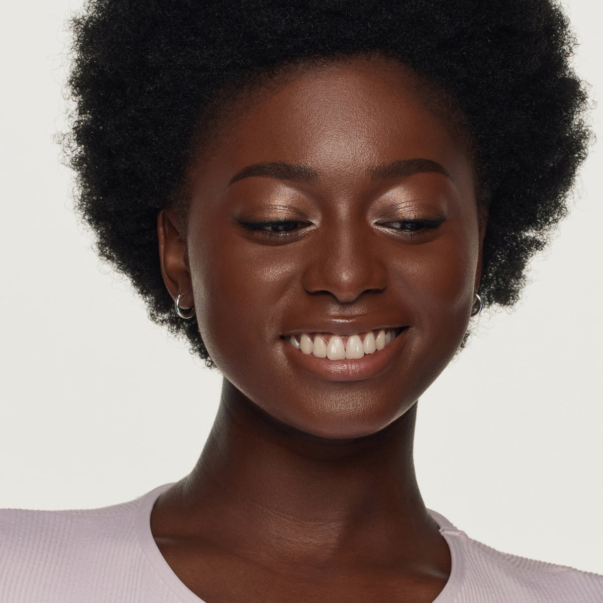 Eine junge Frau mit kurzem Afro lächelt nach unten. Sie trägt dezenten Lidschatten in Nude-Tönen, Mascara und ein helles Oberteil.