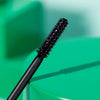 Nahaufnahme des Zauberstabs der Essence lash WITHOUT LIMITS TUBING Extreme Lengthening & Volume Mascara 04 vor einem unscharfen grünen und türkisfarbenen Hintergrund.