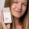 Eine Frau, die lächelt und eine weiße Flasche „Transparent Lab Oil-Control Ultra Fluid Cica Sunscreen SPF 50+“ nahe an ihr Gesicht hält.