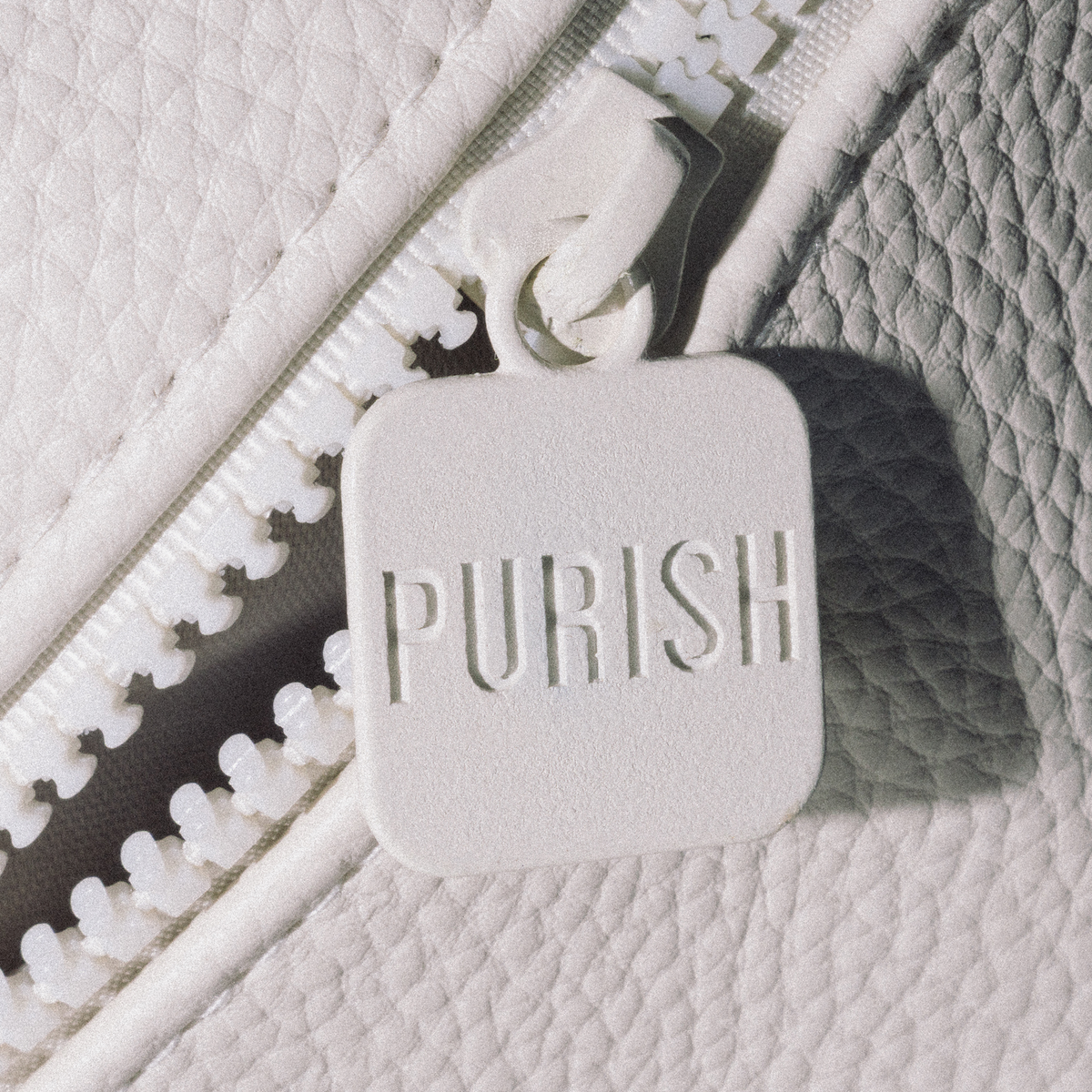 Nahaufnahme der PURISH-Reisetasche – Cremefarben, mit einem weißen Reißverschluss und einem quadratischen Schieber mit der Aufschrift „PURISH“ auf strukturiertem Stoff.