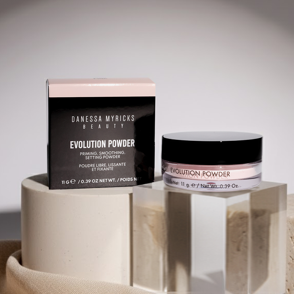 Evolution Powder Pink von Danessa Myricks Beauty mit Schachtel, ausgestellt auf geometrischen Steinplattformen unter sanfter Beleuchtung.