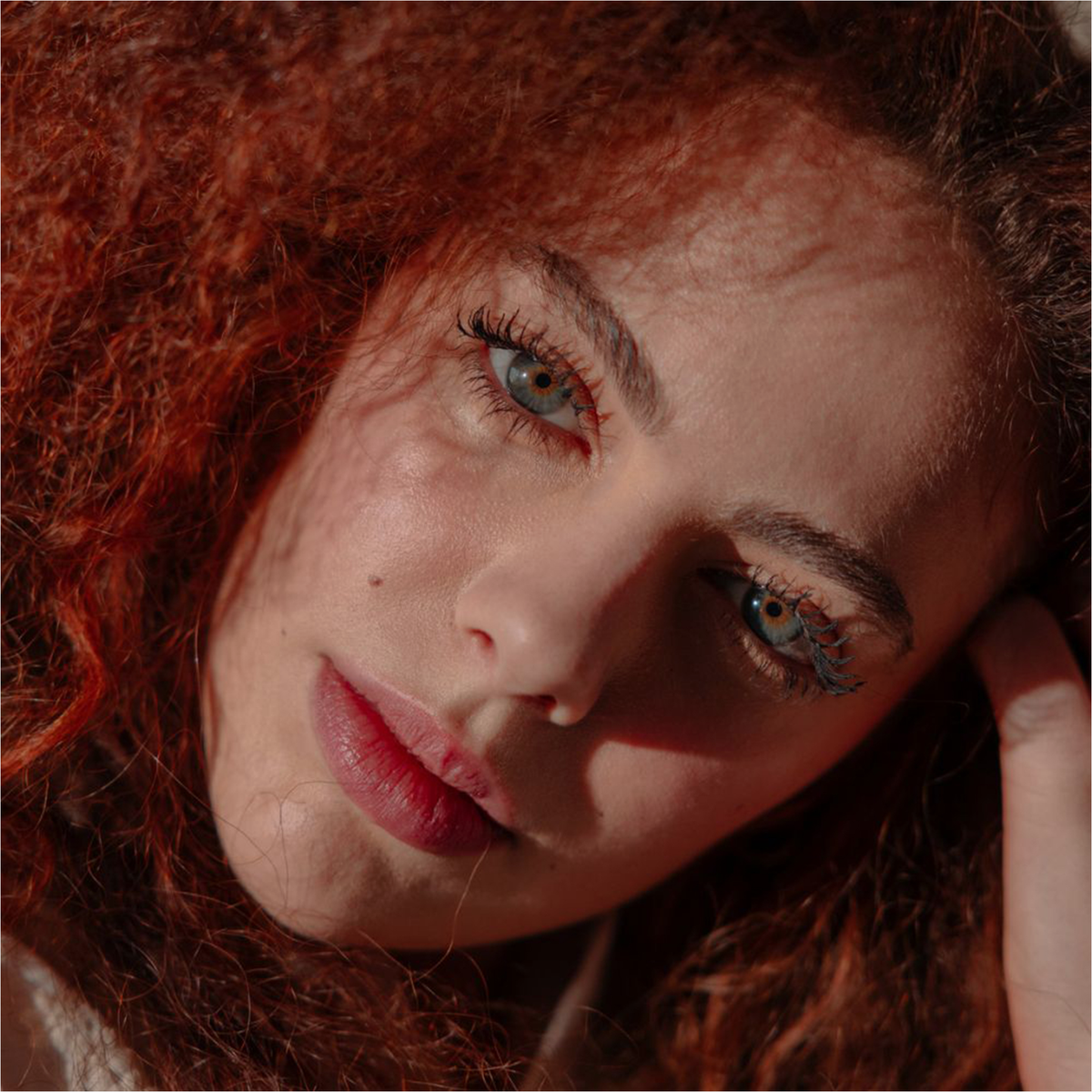 Eine Frau mit lockigem rotem Haar trägt die BLONDESISTER 4 IN 1 CLICK & TURN MASCARA, und das Sonnenlicht lässt ihre blauen Augen strahlen.