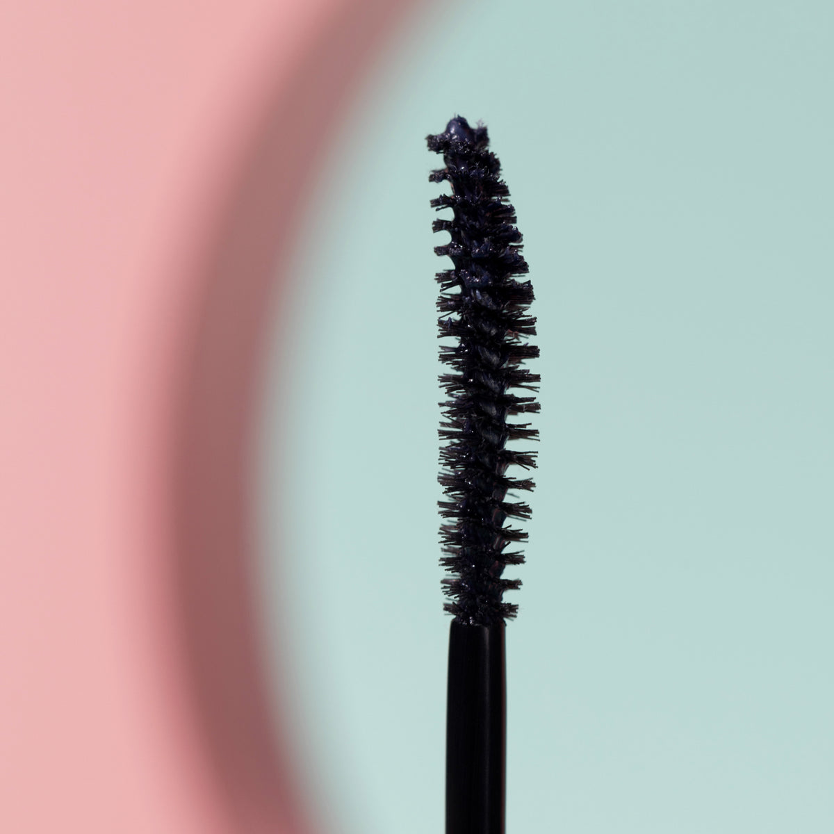 Nahaufnahme des schwarzen, geschwungenen Mascarastifts vor einem Hintergrund in Pastellrosa und Mintgrün.