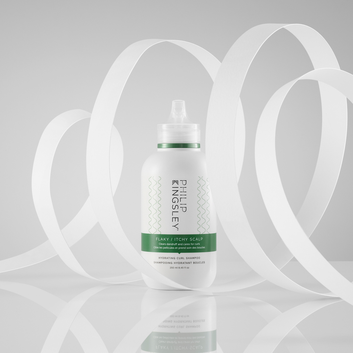 Eine Flasche „Philip Kingsley Flaky/Itchy Scalp Hydrating Curl Anti-Dandruff Shampoo“ steht inmitten weißer Wirbel auf einem hellgrauen Hintergrund.