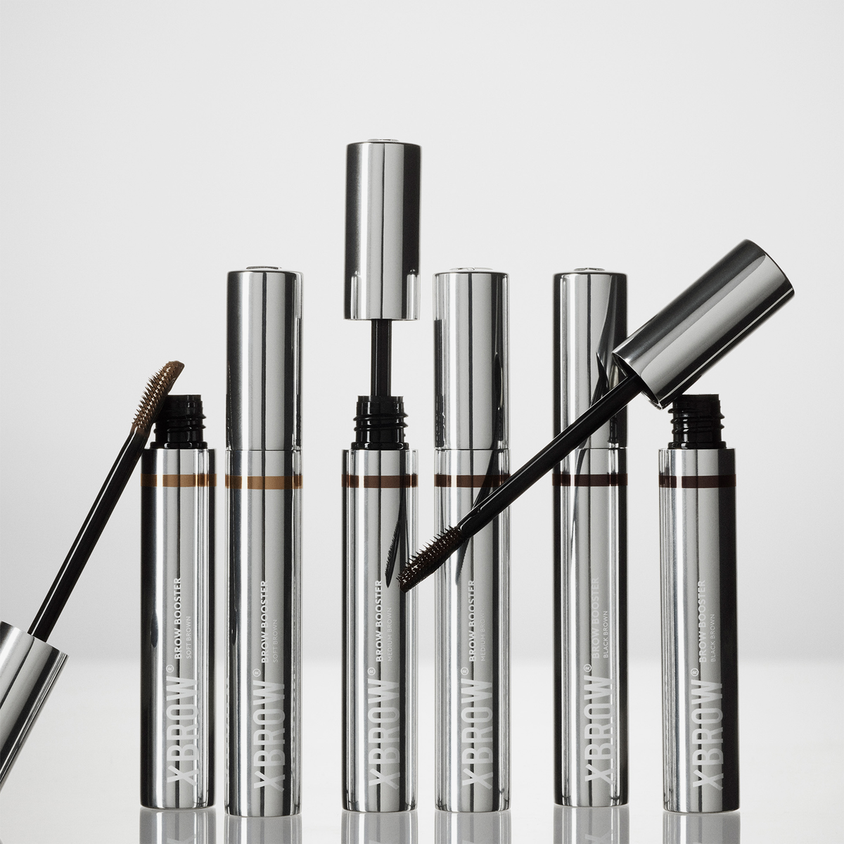 Sechs silberne XLASH Brow Booster-Tuben mit Applikatorstäbchen, von denen einige geöffnet abgebildet sind, zusammen mit Pinseln für getöntes Augenbrauengel.
