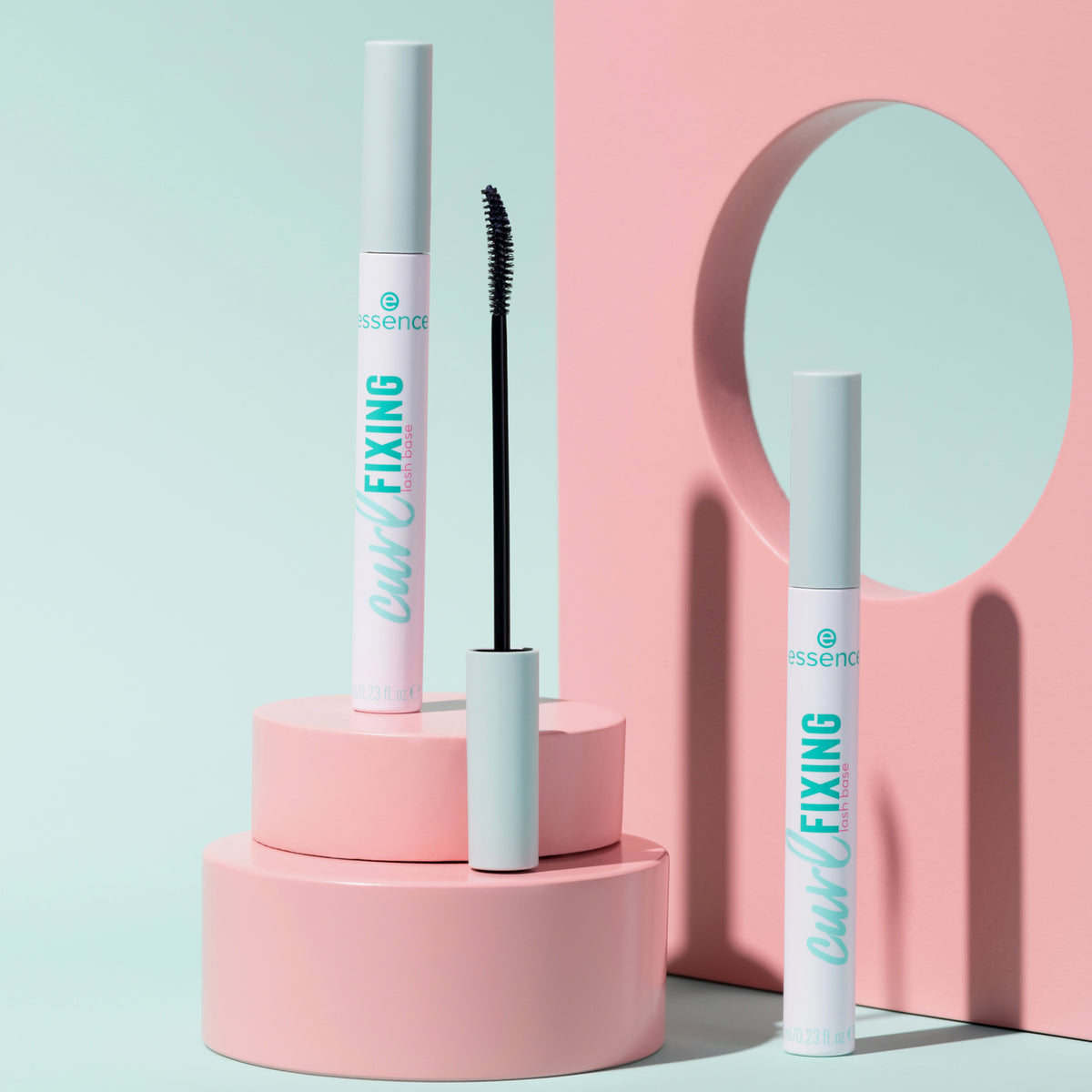 Drei Essence Curl Fixing Lash Base Mascara in Weiß mit gebogenem Bürstchen, auf rosa Podesten vor pastellblauem Hintergrund arrangiert.