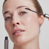 Eine Frau trägt den „XLASH Drama Tinted Primer“ auf ihre oberen Wimpern auf und hält dabei den Applikator dicht an ihr Auge.