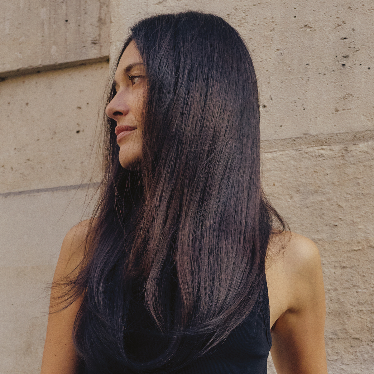 Frau mit langem, glattem, glänzendem dunklem Haar blickt zur Seite vor heller Steinwand.