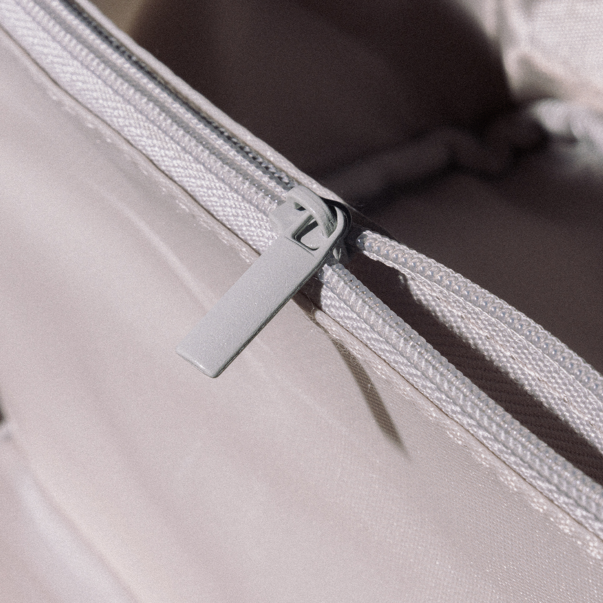 Nahaufnahme eines hellgrauen, teilweise geöffneten Reißverschlusses an der PURISH Reisetasche – Creme, wobei das Sonnenlicht sanfte Schatten wirft.