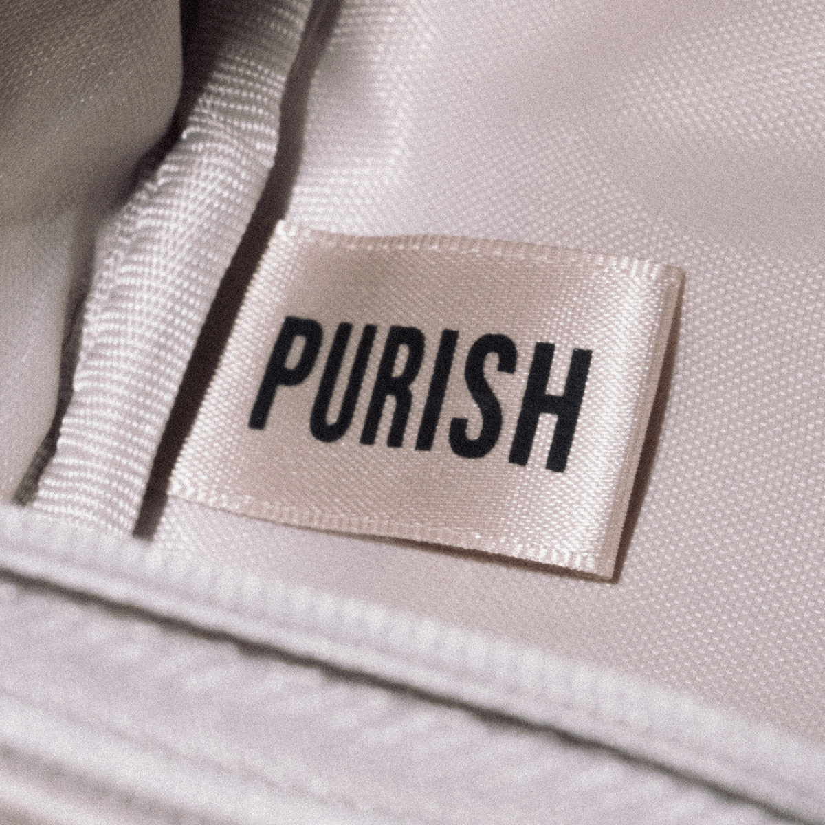 An der cremefarbenen PURISH-Reisetasche ist ein weißes Stoffetikett mit der Aufschrift „PURISH“ in schwarzer Schrift aufgenäht.