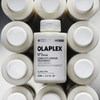 Eine Flasche OLAPLEX N°3 PLUS Complete Repair Treatment steht zwischen identischen Flaschen, von oben betrachtet auf einer silbernen Fläche.