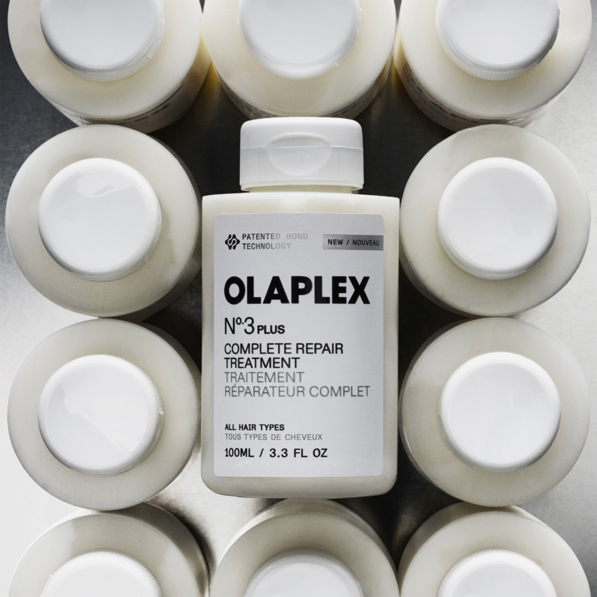 Eine Flasche OLAPLEX N°3 PLUS Complete Repair Treatment steht zwischen identischen Flaschen, von oben betrachtet auf einer silbernen Fläche.