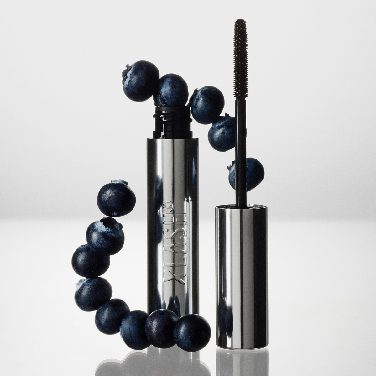 XLASH Lash Booster Mascara-Tube und Applikatorbürste, umgeben von frischen Heidelbeeren.