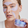 Eine Frau, die lavendelfarbene Longevity Multi-zone Gels unter den Augen und auf der Stirn trägt, während sie ein Töpfchen mit dem Produkt in der Hand hält.
