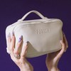 Lange, verzierte Nägel halten die PURISH Purish Favorites Bag – cremefarben mit Struktur und Etikett – vor einem violetten Hintergrund.