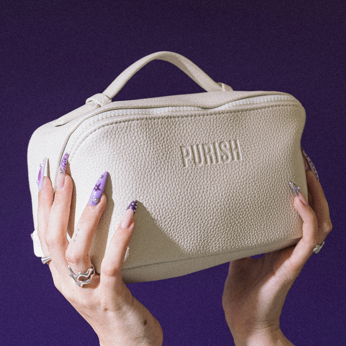 Lange, verzierte Nägel halten die PURISH Purish Favorites Bag – cremefarben mit Struktur und Etikett – vor einem violetten Hintergrund.