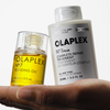 Eine Hand hält die Flaschen OLAPLEX N°3 PLUS Complete Repair Treatment und No.7 Bonding Oil vor einem weißen Hintergrund.
