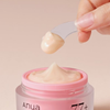 Eine Hand hält einen Spatel mit Creme über einem offenen, rosafarbenen Tiegel mit „ANUA Peach 77 Niacin Enriched Cream“.