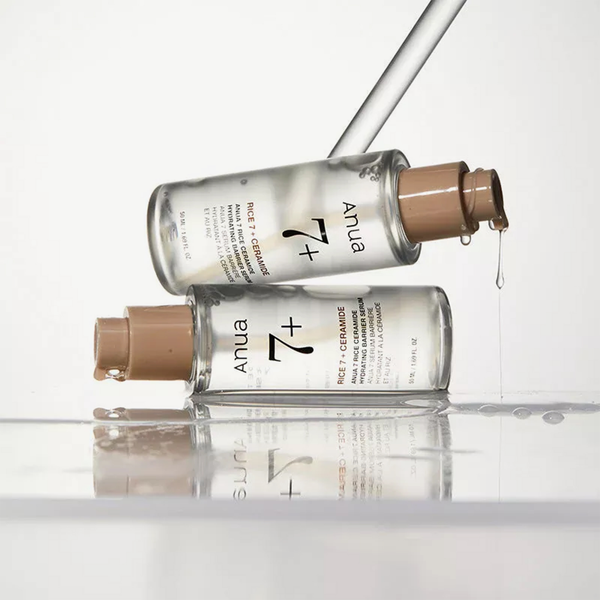 Zwei Flaschen des ANUA Rice Ceramide Hydrating Barrier Serums mit beigen Verschlusskappen, die auf einer nassen Oberfläche übereinander gestapelt sind, tropfen klare Flüssigkeit ab.