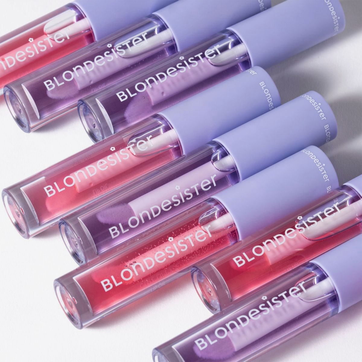 Sieben Tuben „BLONDESISTER 3 IN 1 CUTE LIPPY LIP OIL“ in Rosa und Lila mit lavendelfarbenen Verschlüssen, diagonal auf einer weißen Fläche angeordnet.